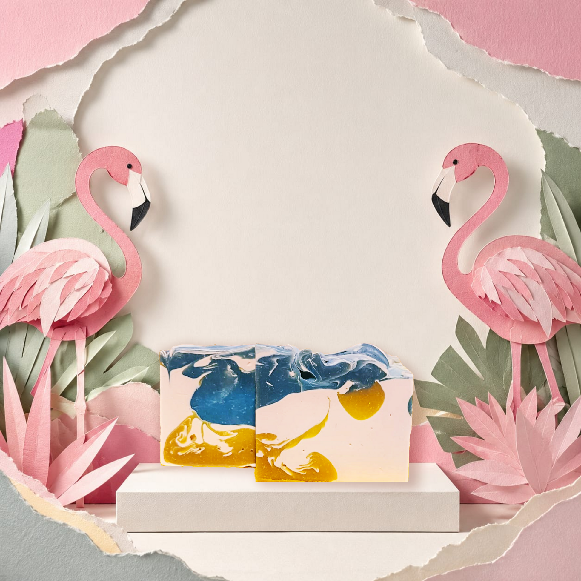 Two rectangular bar soaps with a marbled blue, yellow, and cream design are displayed on a white pedestal. The scene is styled with pink paper-cut flamingos and tropical leaves against a torn-paper backdrop. Mai Tai Artisan SOap