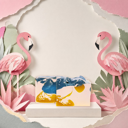 Two rectangular bar soaps with a marbled blue, yellow, and cream design are displayed on a white pedestal. The scene is styled with pink paper-cut flamingos and tropical leaves against a torn-paper backdrop. Mai Tai Artisan SOap